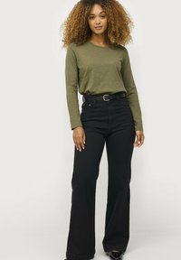 Frau mit lockigem Haar, die ein olivgrünes Langarmshirt, hochgeschnittene schwarze Schlaghosen, einen schwarzen Gürtel und schwarze Schuhe trägt, steht.