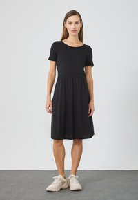 Schwarzes Kurzarmkleid mit Rundhalsausschnitt, geraffter Taille und ausgestellt Rock. Model kombiniert mit hellen Sneakers. Glatte Textur.