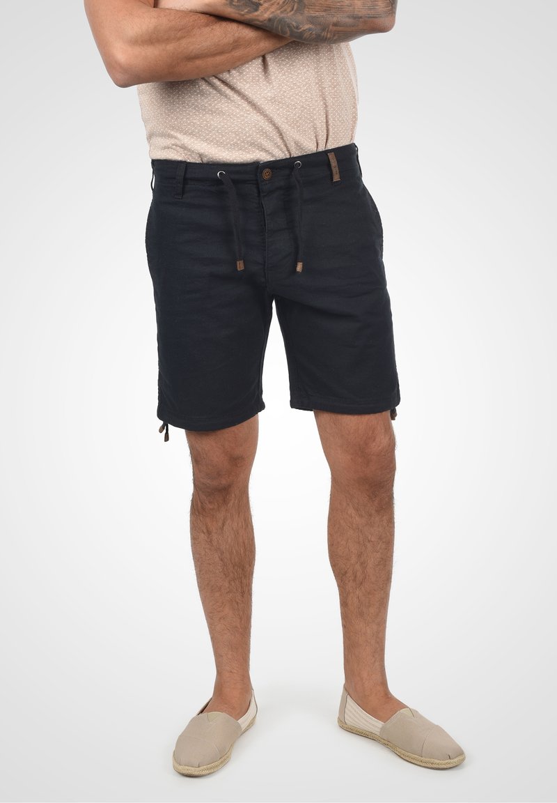 Shorts noirs avec une taille à cordon de serrage et des accents noués sur les côtés, confectionnés dans un tissu texturé. Associés à des chaussures beiges à enfiler.