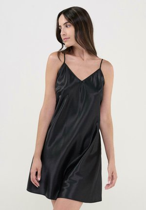 Mujer con cabello largo y oscuro, vistiendo un vestido corto de raso negro sin mangas, mirando hacia su izquierda contra un fondo claro y sencillo.