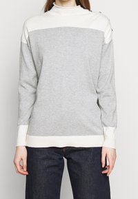 Grå tröja med hög hals, med vita horisontella ränder, ribbade muddar och skålad knappdetalj på axlarna. Mjuk stickad tyg, avslappnad passform.