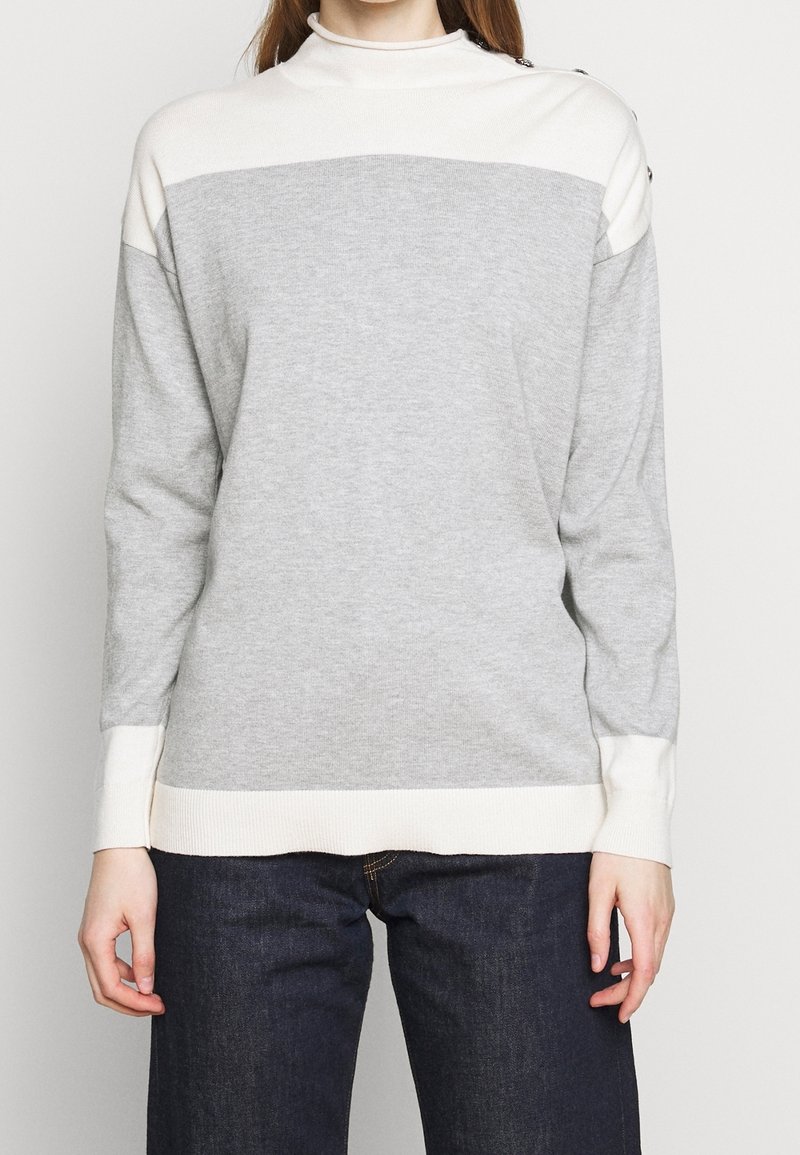 Grå tröja med hög hals, med vita horisontella ränder, ribbade muddar och skålad knappdetalj på axlarna. Mjuk stickad tyg, avslappnad passform.