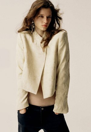 Jonge vrouw die een crème textuur cropped jack draagt, zwarte broek, lang golvend haar en grote zilveren hangende oorbellen, staand met handen in de zakken.