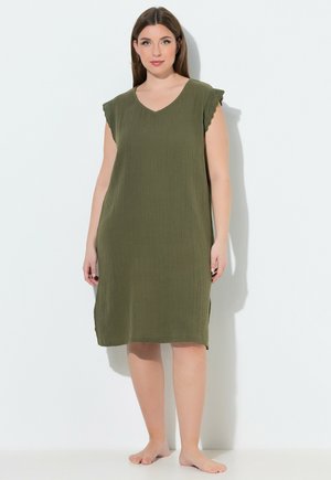 Frau in einem ärmellosen, olivgrünen knielangen Kleid mit welligen Spitzenärmeln, barfuß stehend vor weißem Hintergrund.