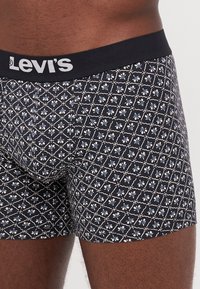 Crne bokserice s geometrijskim bijelim i svijetloplavim cvjetnim uzorkom, s crnim Levi's pojasom. Mekan, prilagođen dizajn.