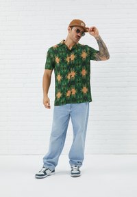 Kortermet grønn skjorte med et flerfarget edderkoppmønster, kombinert med lyseblå jeans og mørke sneakers. Tilbehør inkluderer solbriller og en caps.