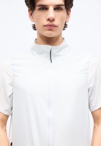 Νεαρός άνδρας με κοντά καστανά μαλλιά που φοράει λευκό αμάνικο γιλέκο με φερμουάρ πάνω από λευκή κοντομάνικη μπλούζα, μπροστά σε λευκό φόντο.