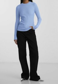 Frau trägt einen hellblauen, gerippten Pullover mit langen Ärmeln, schwarze Hosen mit weitem Bein und metallisch spitz zulaufende Schuhe, steht vor einem schlichten Hintergrund.