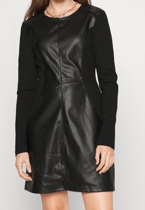 Frau trägt ein eng anliegendes schwarzes Kleid mit langen Ärmeln und einem vorderen Paneel aus glänzendem, lederähnlichem Material.