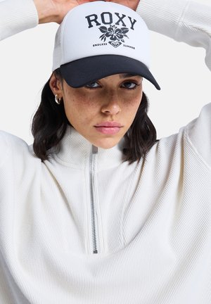 Biela čiapka Roxy s čiernym šiltom a logom s kvetinovým motívom. Modelka má na sebe textúrovaný biely top so zipsom na krku. Vlasy sú čiastočne zasunuté za uši.