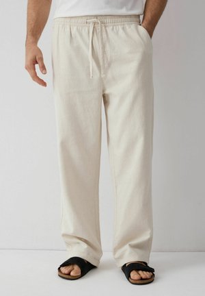 Mężczyzna noszący luźne beżowe spodnie ze sznurkiem i czarne sandały, stojący na jednolitym białym tle.