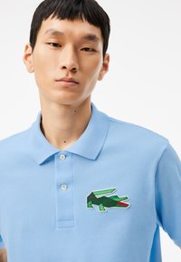 Ljusblå polo skjorta i bomull, med en texturerad tyg och en grön, svart och röd broderad krokodil-logotyp på bröstet.