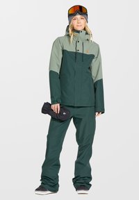 Grüne und graue Outdoor-Jacke mit passender Hose, aus wasserdichtem Material gefertigt. Enthält eine Kapuze, Reißverschlusstaschen und wird mit Skihandschuhen kombiniert.