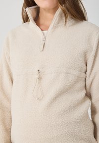 Bež villane pullover kõrge kraega, pooltõmbluku ees ja reguleeritava nööriga vööl. Pehme, tekstuuriga kangas.