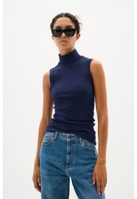 Frau trägt ärmelloses marineblaues Rollkragentop, blaue Jeans und große schwarze Sonnenbrille, steht vor einem schlichten hellen Hintergrund.