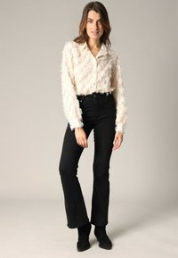 Vrouw in een crèmekleurige, gestructureerde blouse met lange mouwen, zwarte hoge taille jeans en zwarte enkellaarsjes, staande tegen een effen achtergrond.