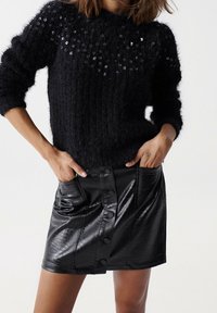 Černý huňatý pletený svetr s flitrovým designem na výstřihu. Černá mini sukně z umělé kůže s detaily na knoflících a texturovaným povrchem.
