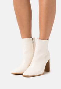Botines de cuero blanco con punta afilada y tacón cuadrado texturizado en marrón; cuentan con una cremallera lateral para poner y quitar fácilmente.