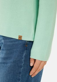 Miętowy sweter z dzianiny o teksturowanej powierzchni, w luźnym kroju, z brązową metką logo na dolnym końcu, w połączeniu z niebieskimi dżinsami.