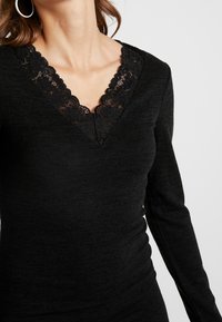 Černý top s dlouhými rukávy, který má hluboký výstřih do V a složitou krajkovou výzdobu podél výstřihu. Tkanina se zdá být měkká a má přiléhavý siluetu.