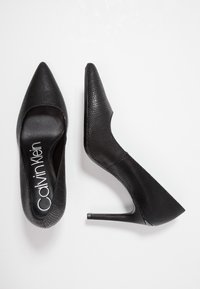 Černé kožené vysoké podpatky s texturovaným povrchem, špičatou špičkou a štíhlým stiletto podpatkem. Logo "Calvin Klein" viditelné na stélce.