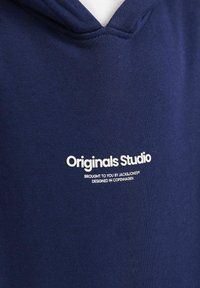 Mornarsko plava dukserica s kapuljačom izrađena od mekanog materijala. Ima bijeli otisnuti logo: "Originals Studio" i dodatni tekst ispod loga.