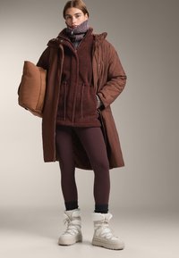 Hnědý prošívaný dlouhý kabát přes bordó fleecovou mikinu a hnědé legíny. Modelka má na nohou bílé zateplené boty se šedými doplňky a drží hnědý polštář.