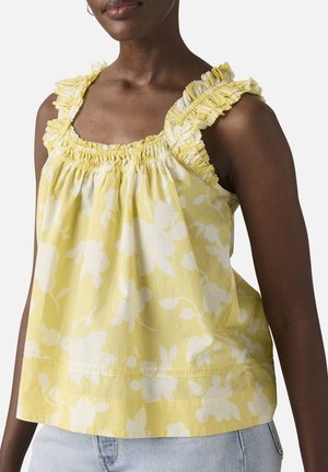 Femme portant un haut jaune sans manches avec des motifs floraux blancs et des bretelles à volants, associé à un jean bleu clair.