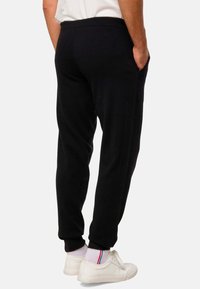 Pantalon de survêtement noir ajusté, avec poches latérales et poignets élastiques ; associé à des baskets blanches et des chaussettes avec des rayures rouges et bleues.