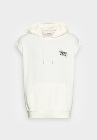 Ljus beige ärmlös hoodie gjord av mjukt tyg; har en framficka, dragsnoddar och broderad text med blommönster.