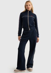 Survêtement bleu marine avec une veste zippée et un pantalon large, présentant un passepoil crème et des accents de logo rouges. Tissu doux et texturé avec un design sportif.