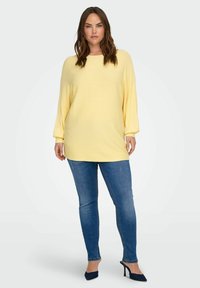 Geltona riboti megzta tunika su ilgomis rankovėmis, derinama su mėlynais siaurais džinsais ir tamsiai mėlynais aukštakulniais, turi apvalų iškirptę ir laisvą siluetą.