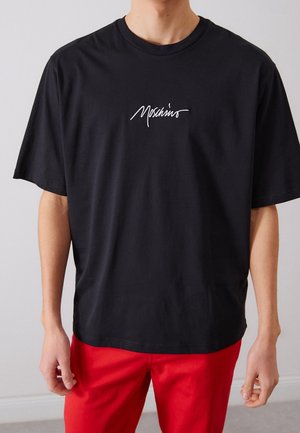 Henkilö, jolla on musta Moschinon t-paita ja punaiset housut, seisoo yksivärisen vaalean taustan edessä.