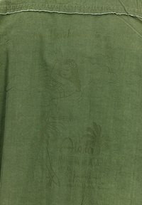 T-shirt in tessuto verde con un design grafico che presenta una figura, elementi tropicali e testo in una tonalità di verde più chiara; orlo leggermente consumato.