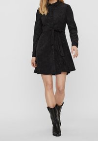 Schwarzes Wildlederhemdkleid mit langen Ärmeln, Knopfleiste, Gürtel in der Taille, einem ausgestellten Rock und getragen mit kniehohen schwarzen Stiefeln.
