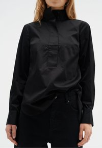 Schwarzes Langarmshirt mit hohem Kragen, das mit einer Knopfleiste und glatter Textur ausgestattet ist. Kombination mit dunklen Jeans, die ein minimalistisches Design betonen.