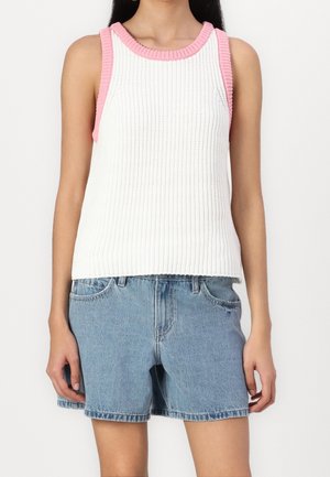 Femme portant un haut sans manches en maille blanche avec des bordures roses et un short en denim bleu clair sur un fond uni.