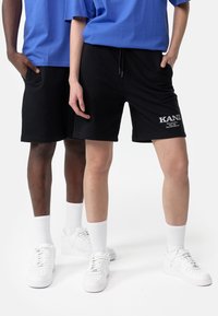 Černé bavlněné kraťasy s tkaničkou, s bílým nápisem značky "KANI". Nosené s bílými ponožkami a teniskami.