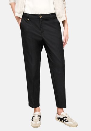 Frau trägt schwarze, maßgeschneiderte, knöchellange Hosen, weiße und schwarze Turnschuhe sowie eine cremefarbene, gerippte Jacke über einem weißen Oberteil.