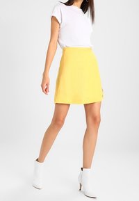 Gelber Rock aus leichtem Stoff, mit ausgestelltem Design, kombiniert mit einem weißen T-Shirt und weißen Stiefeletten. Schlichte Hardware-Details.