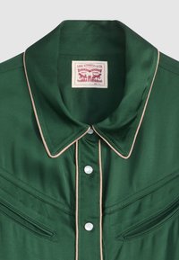 Grünes Satinhemd mit spitzem Kragen, kontrastierendem beigem Piping, Perlmutt-Druckknöpfen und zwei vorderen Taschen mit gesteppten Details. Markenetikett sichtbar.