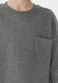 Šedá mikina vyrobená z jemného materiálu, s kulatým výstřihem, malou kapsou na levém prsu a pružnými manžetami a lemem.