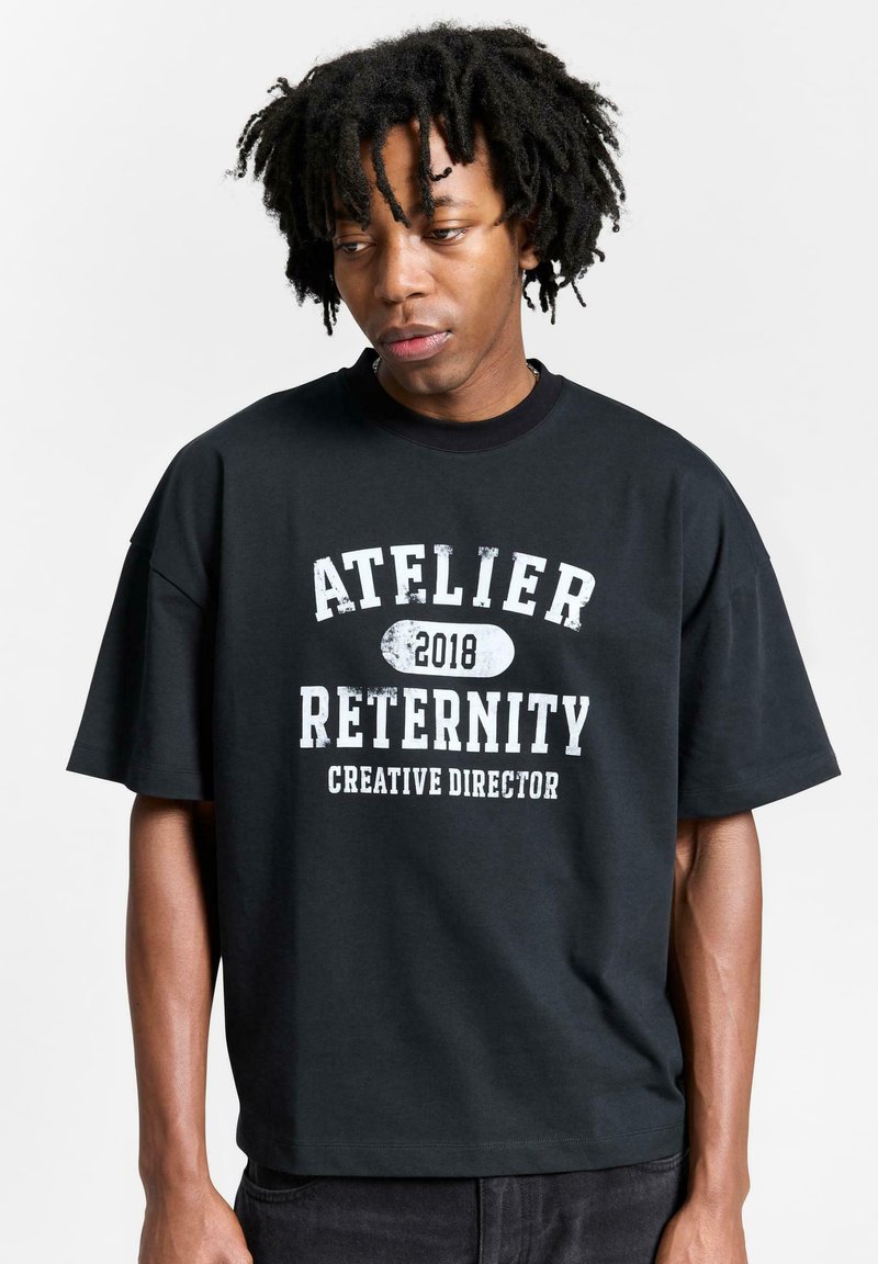 Sort oversized t-shirt lavet af bomuld med hvid trykt tekst: "ATELIER RETERNITY 2018 CREATIVE DIRECTOR." Korte ærmer og afslappet pasform.