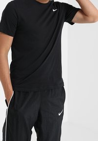 Muž oblečený v černém tričku Nike a černých sportovních kalhotách Nike s bílými pruhy, stojící se zvednutou jednou rukou a druhou rukou v kapse.
