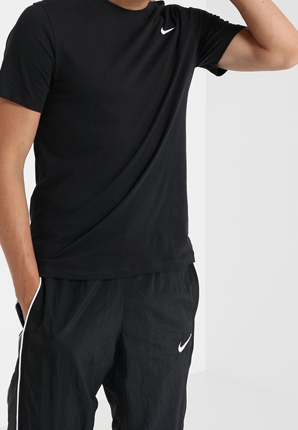 Muž oblečený v černém tričku Nike a černých sportovních kalhotách Nike s bílými pruhy, stojící se zvednutou jednou rukou a druhou rukou v kapse.