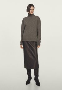 Frau in einem braunen Rollkragenpullover, einem dunkelbraunen Leder-Midikleid und schwarzen knielangen Stiefeln, die vor einem schlichten Hintergrund steht.