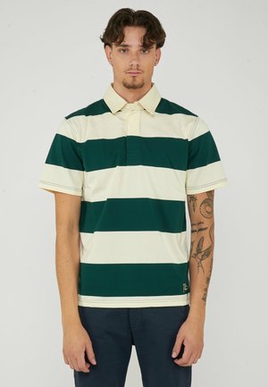 Giovane uomo con capelli corti e scuri che indossa una polo a righe verdi e bianche a maniche corte e pantaloni scuri, con tatuaggi sul braccio sinistro.