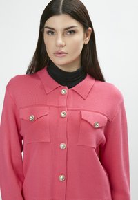 Cárdigan de punto rosa con cuello, que cuenta con dos bolsillos frontales y botones brillantes en tono dorado. Textura suave y ajuste entallado.