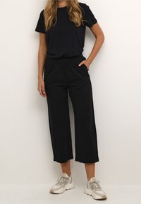 Černé tričko s krátkým rukávem a černé volné kalhoty zkrácené nad kotníky s elastickým pasem a bočními kapsami, doplněné bílými a šedými teniskami.