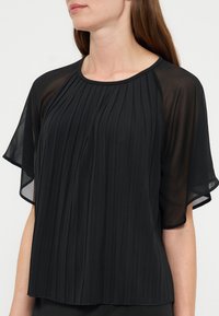 Femme portant un chemisier noir plissé avec des manches courtes transparentes et amples et un col rond.